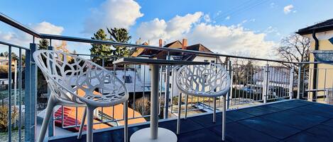 A balcony with two white chairs and a table, overlooking a residential area with houses and trees.