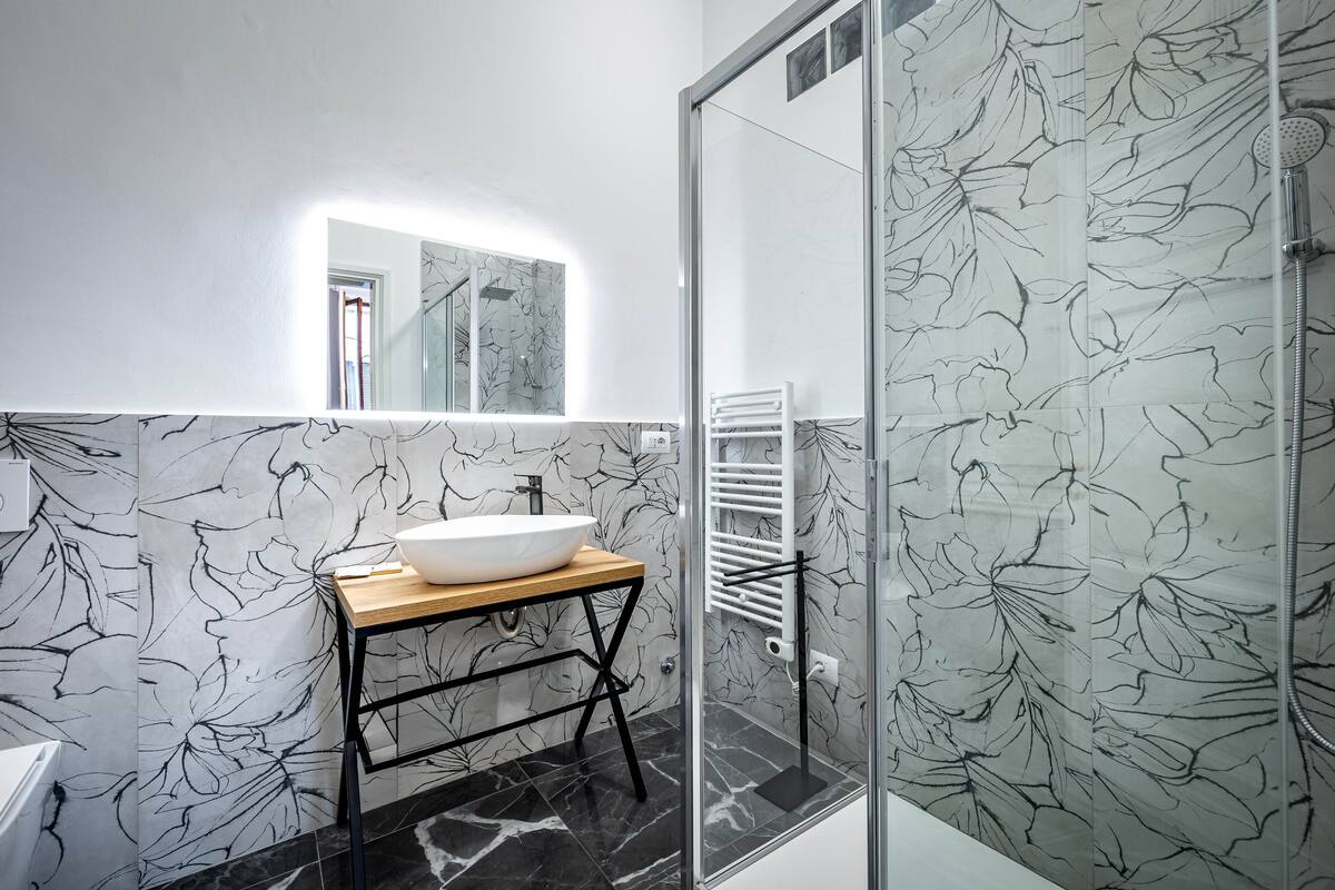 A modern bathroom with a large mirror, a vessel sink, a glass shower enclosure, and a marble-patterned wall.