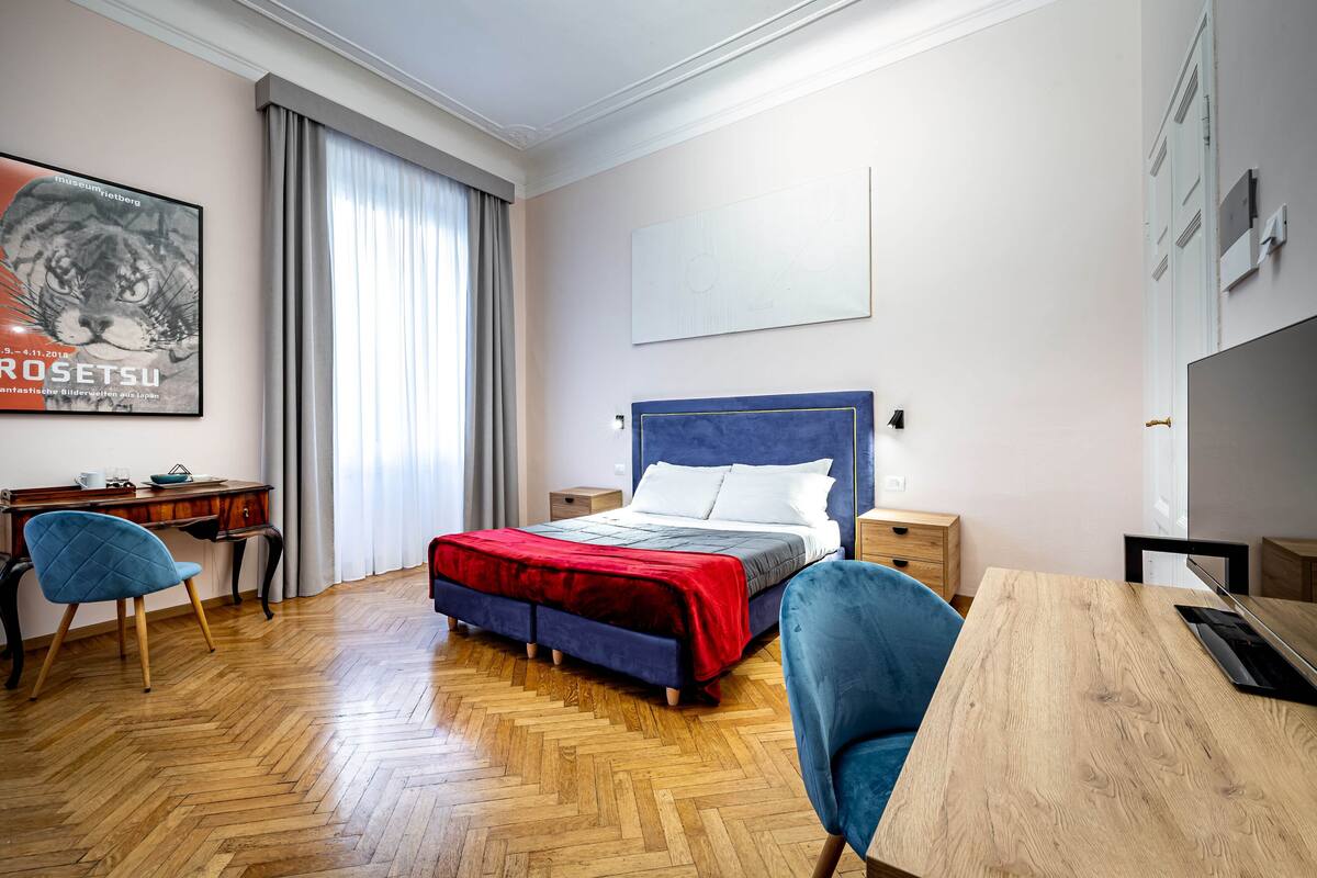 A hotel room with a bed, a desk, a chair, and a wooden floor.