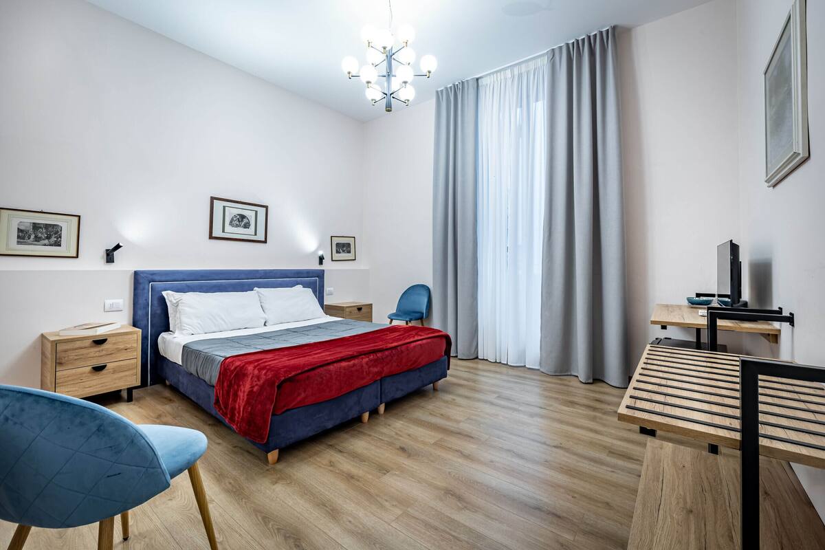 A bedroom with a bed, bedside tables, a desk, a chair, and a large window with curtains.