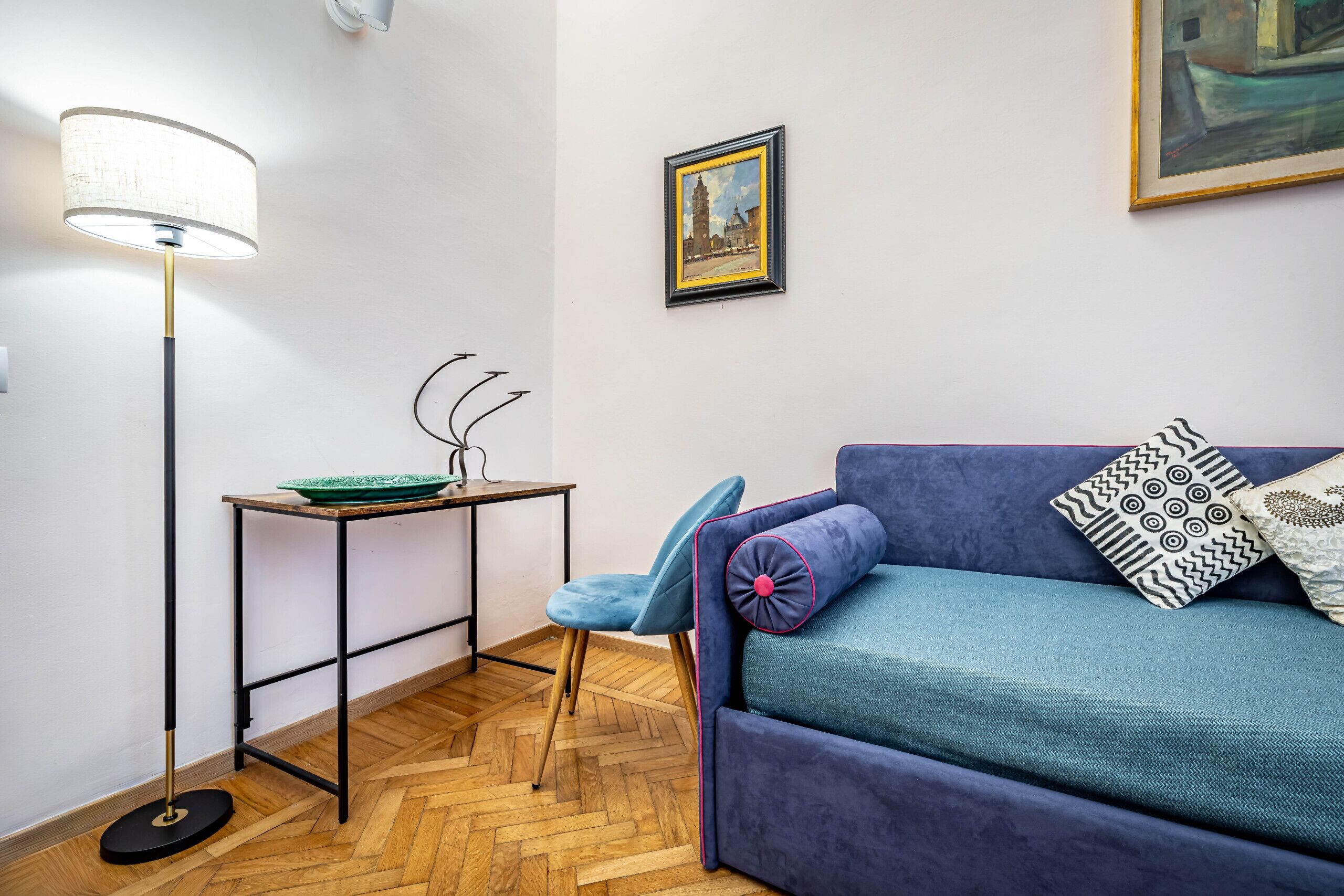 A room with a blue sofa, a floor lamp, a small table with a green tray, and a framed painting on the wall.