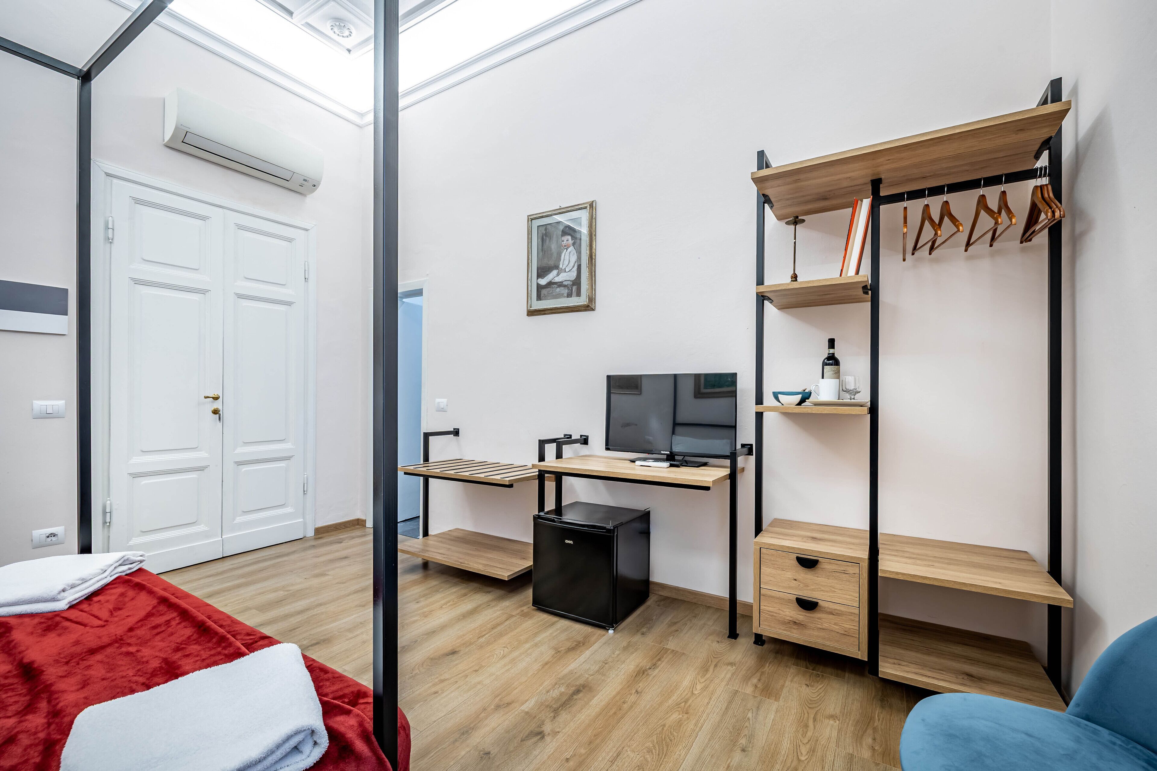 A compact room with a desk, a computer, a small fridge, a wardrobe, and a bed with a red blanket.