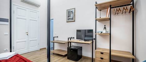 A compact room with a desk, a computer, a small fridge, a wardrobe, and a bed with a red blanket.