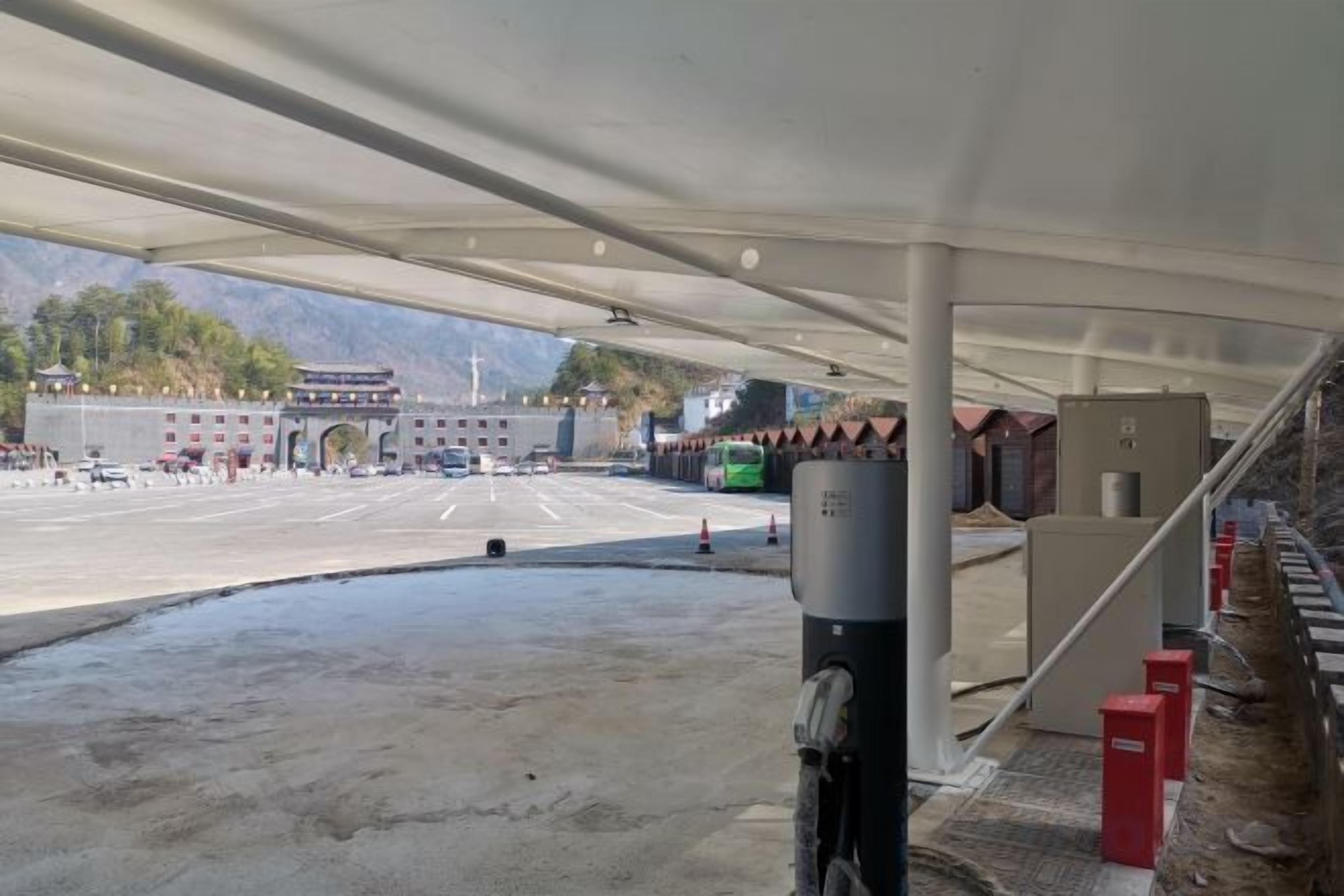 Un área de estacionamiento techada con un edificio grande al fondo, un autobús verde y una barrera roja.