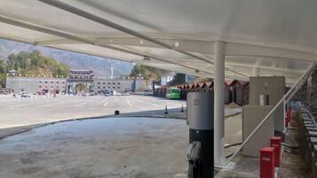 Un área de estacionamiento techada con un edificio grande al fondo, un autobús verde y una barrera roja.