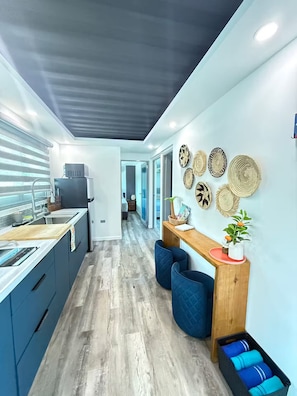 A modern kitchen with a central island, a dining area with a wooden table and blue chairs, and a hallway leading to other rooms. - The Palms Container House (San Antonio)