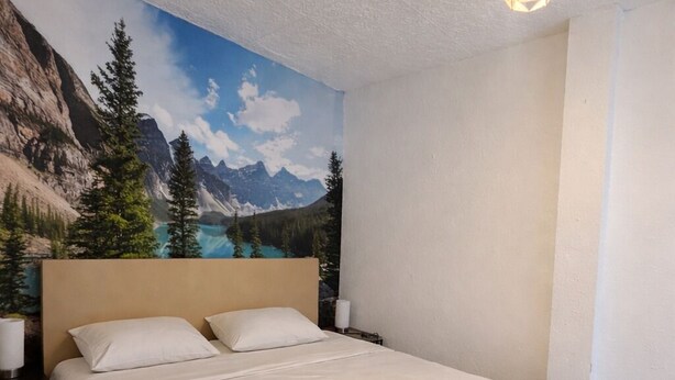 A hotel room with a bed, a nightstand, a lamp, and a mural of a mountain landscape.