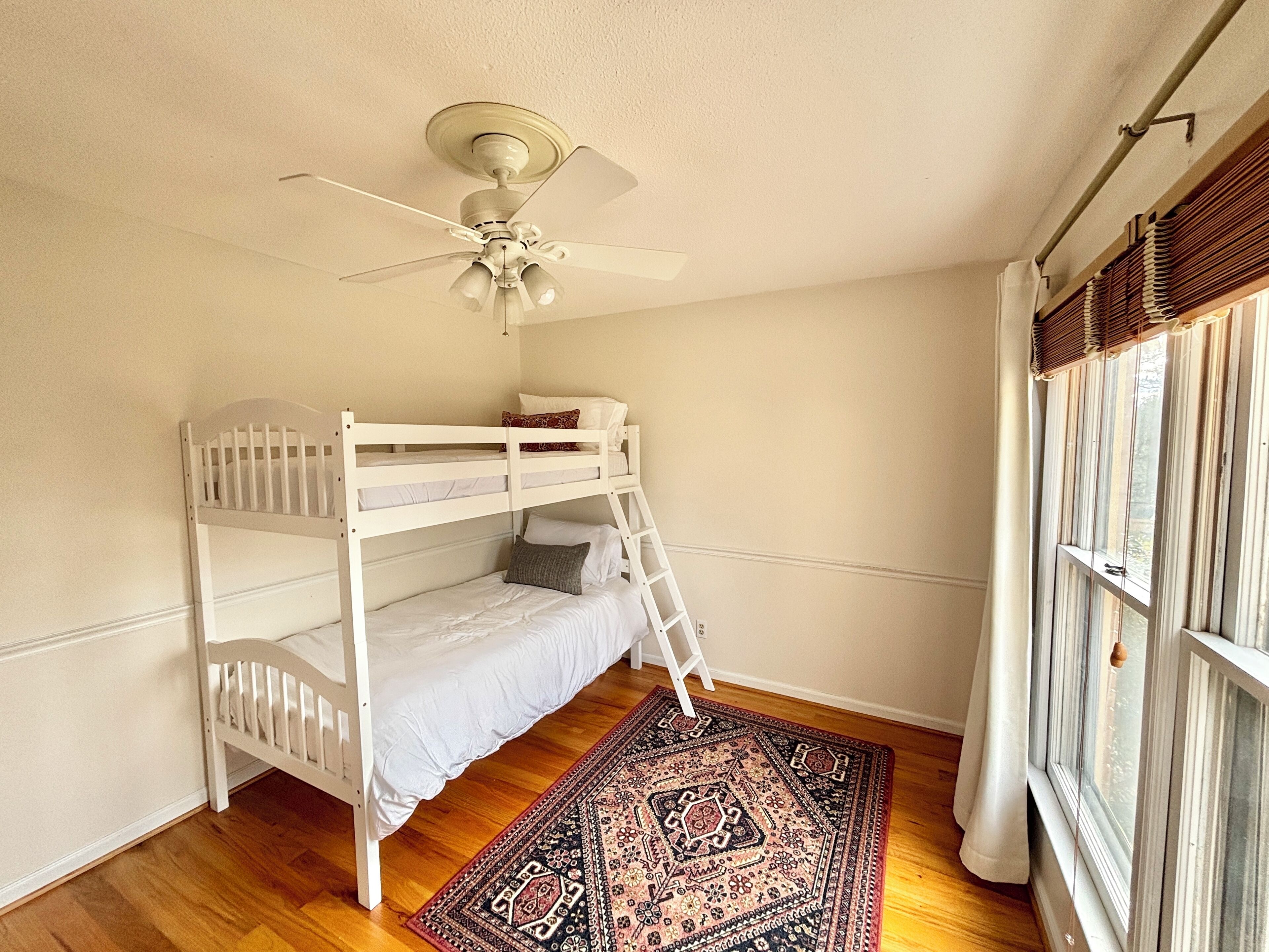 Quarto com beliche, ventoinha no teto, tapete estampado e janela com cortinas.