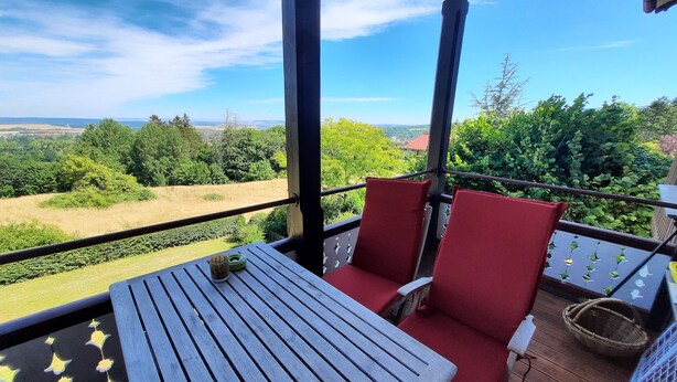 Un balcone con tavolo e due sedie rosse, affacciato su un paesaggio suggestivo con alberi e campi.