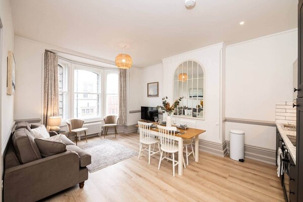 A modern living room with a sofa, a dining table with chairs, a TV, and a large window with curtains. - Grade II Listed 2 Bed Flat in Central Salisbury (Salisbury)