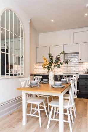 A dining area with a wooden table and white chairs, a vase with flowers, and a kitchen in the background. - Grade II Listed 2 Bed Flat in Central Salisbury (Salisbury)