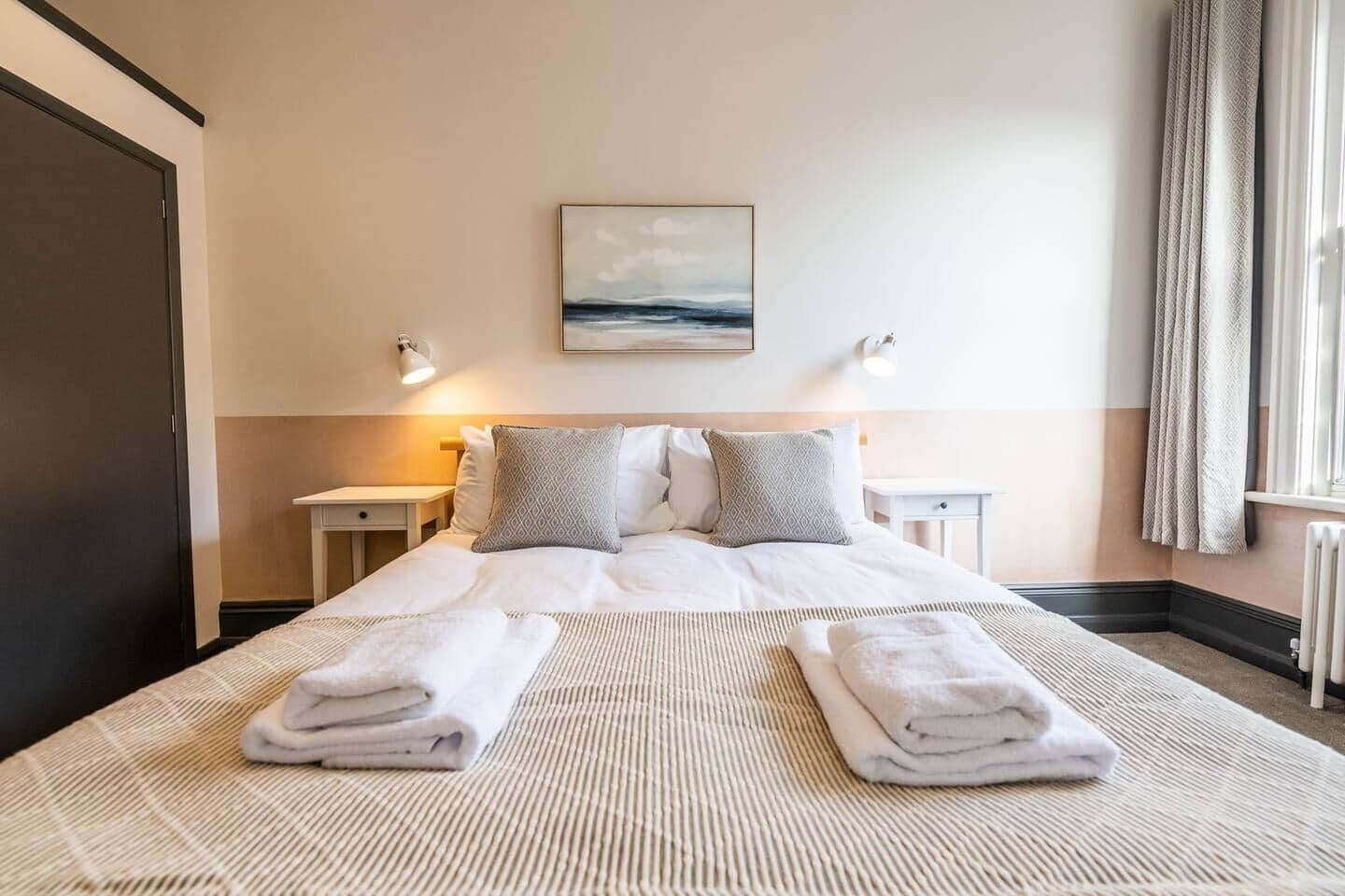 A neatly made bed with two pillows, a bedside table with a lamp, and a painting on the wall.