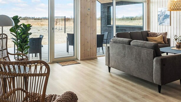 A modern interior with a grey sofa, wooden flooring, and large windows offering a view of the outdoors. - 5 Star Holiday Home in Idestrup (Idestrup)