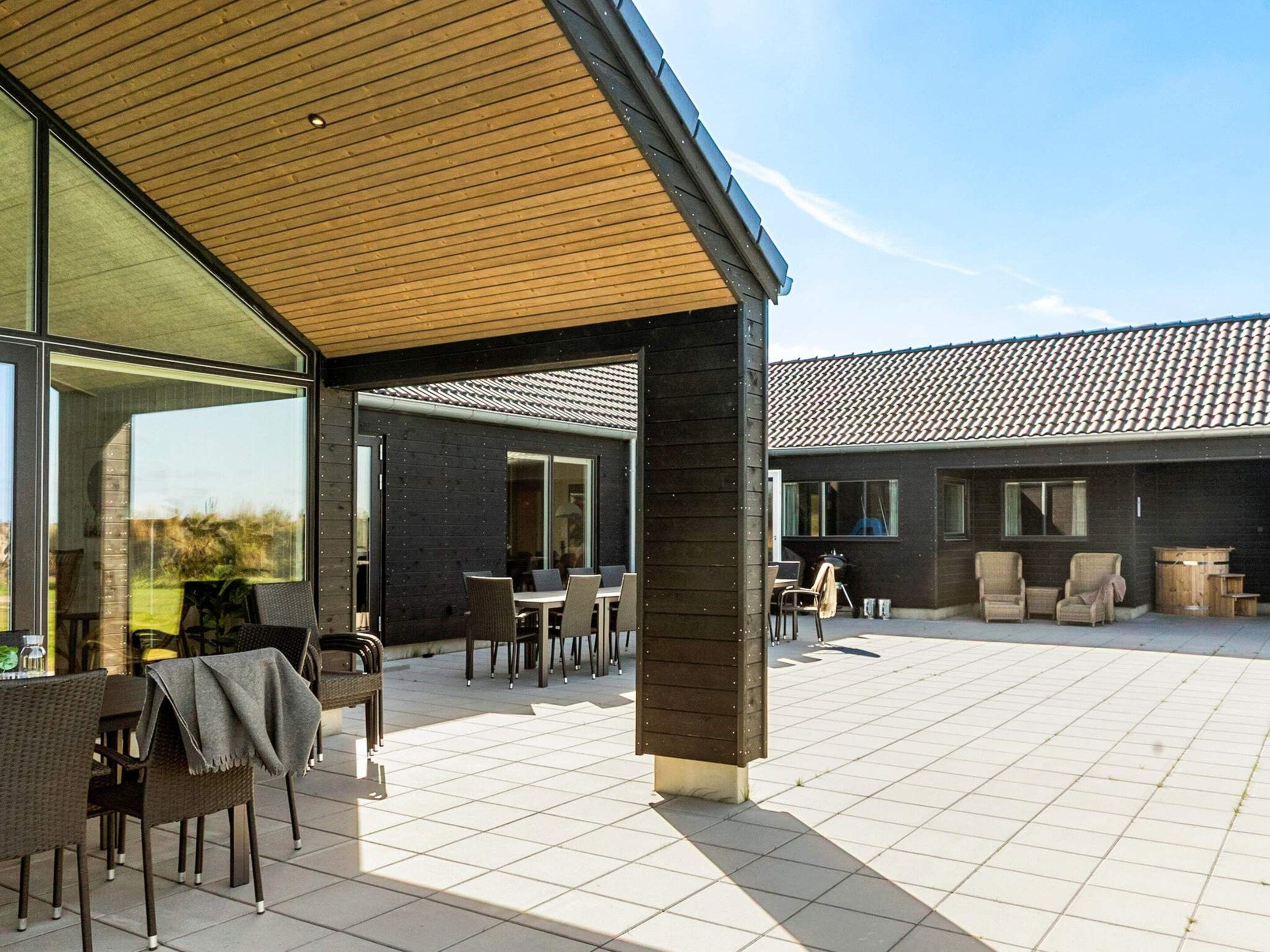 A covered outdoor patio area with seating and tables, a large window, and a view of a building with a tiled roof.