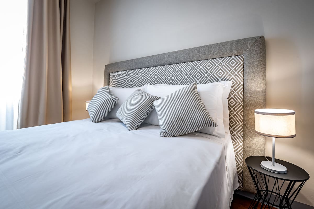 A neatly made bed with white bedding and striped pillows, a patterned headboard, and a small table with a lamp beside it.