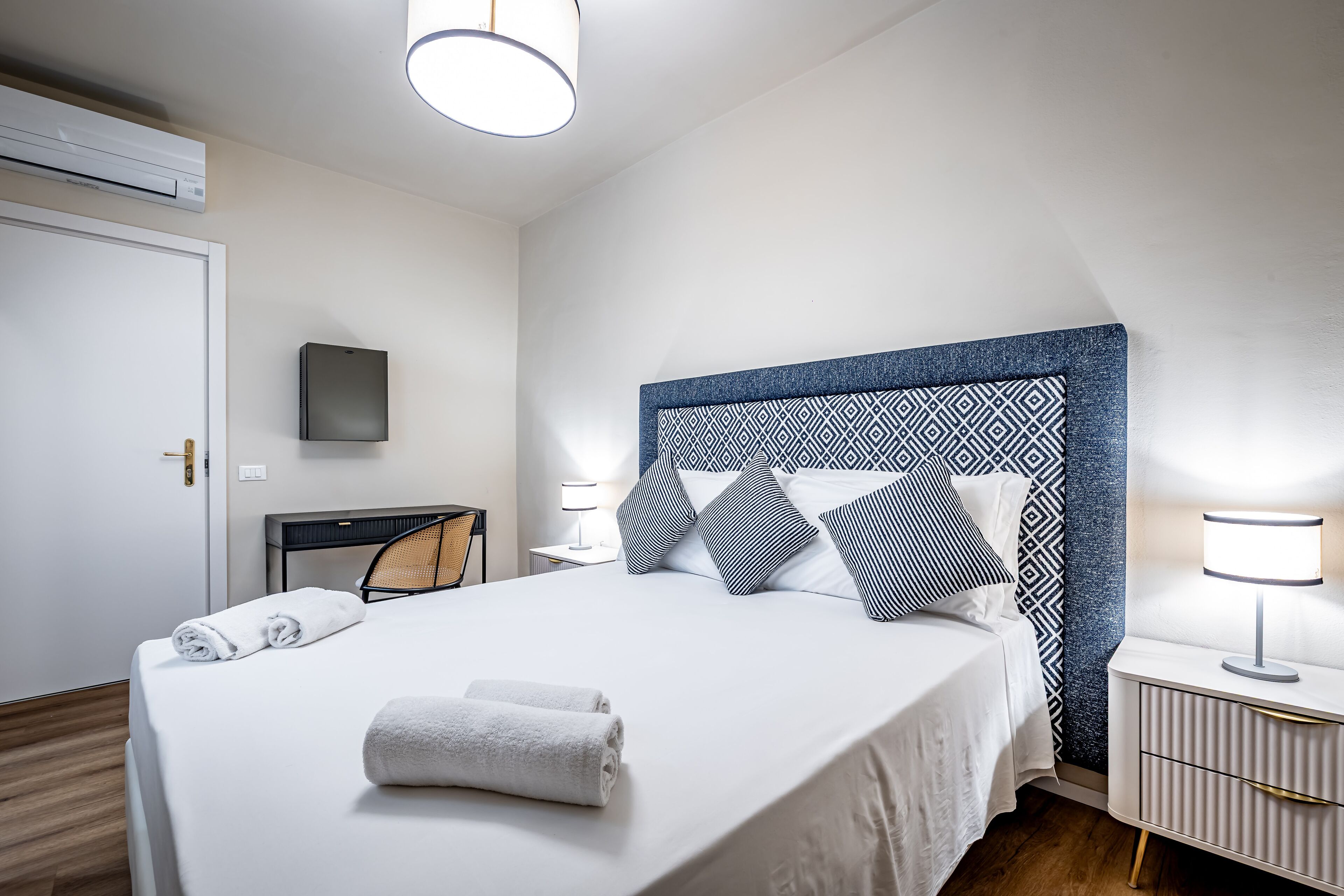 A neatly made bed with white linens and rolled towels, a modern headboard with geometric patterns, a bedside table with a lamp, and a wall-mounted TV.