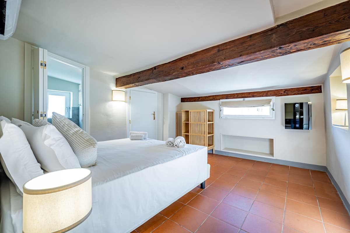 A bedroom with a large bed, a wooden ceiling beam, a built-in wardrobe, and a window.