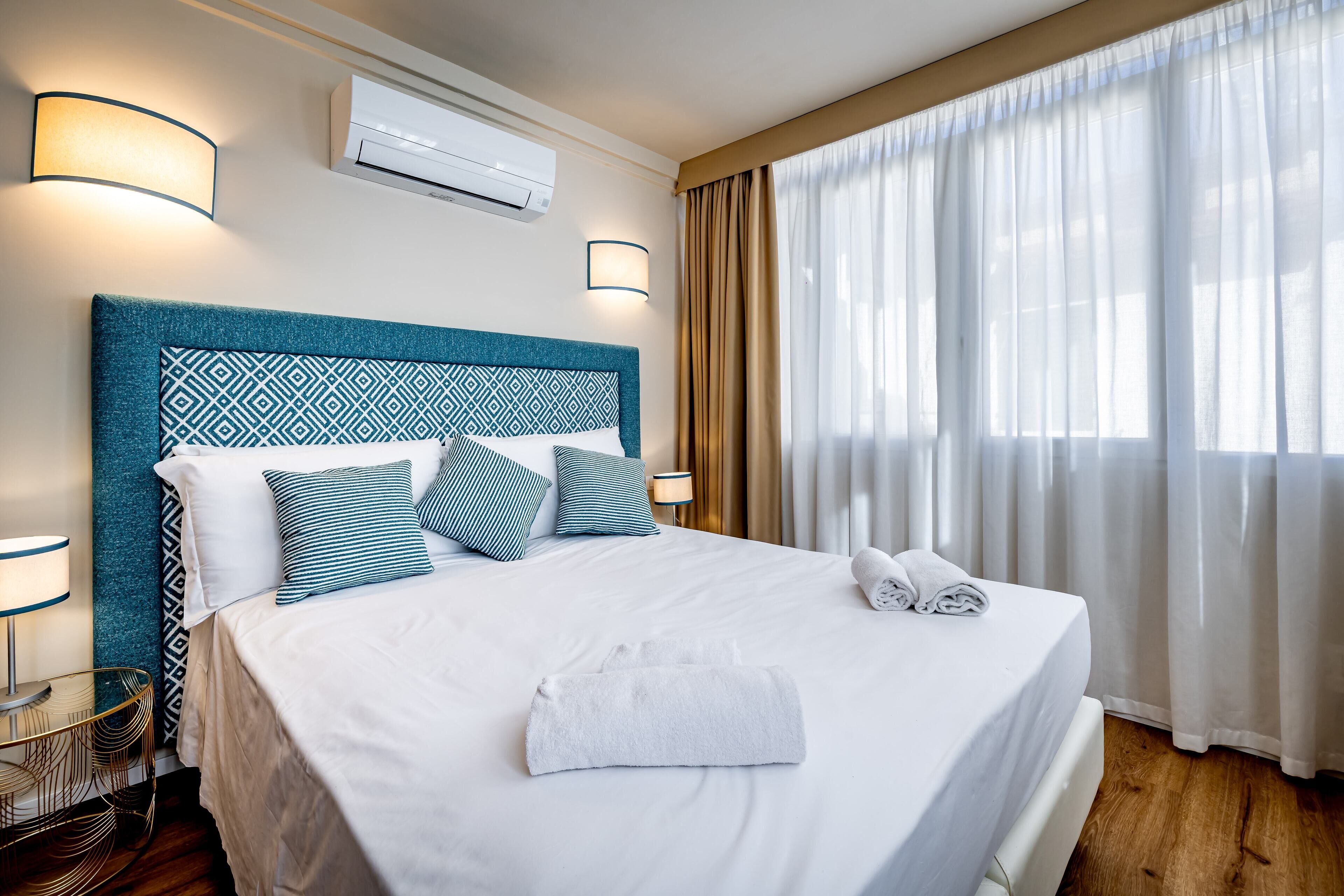Habitación de hotel con una cama grande, cabecera con diseño geométrico, aire acondicionado de pared y ventana con cortinas de gasa.