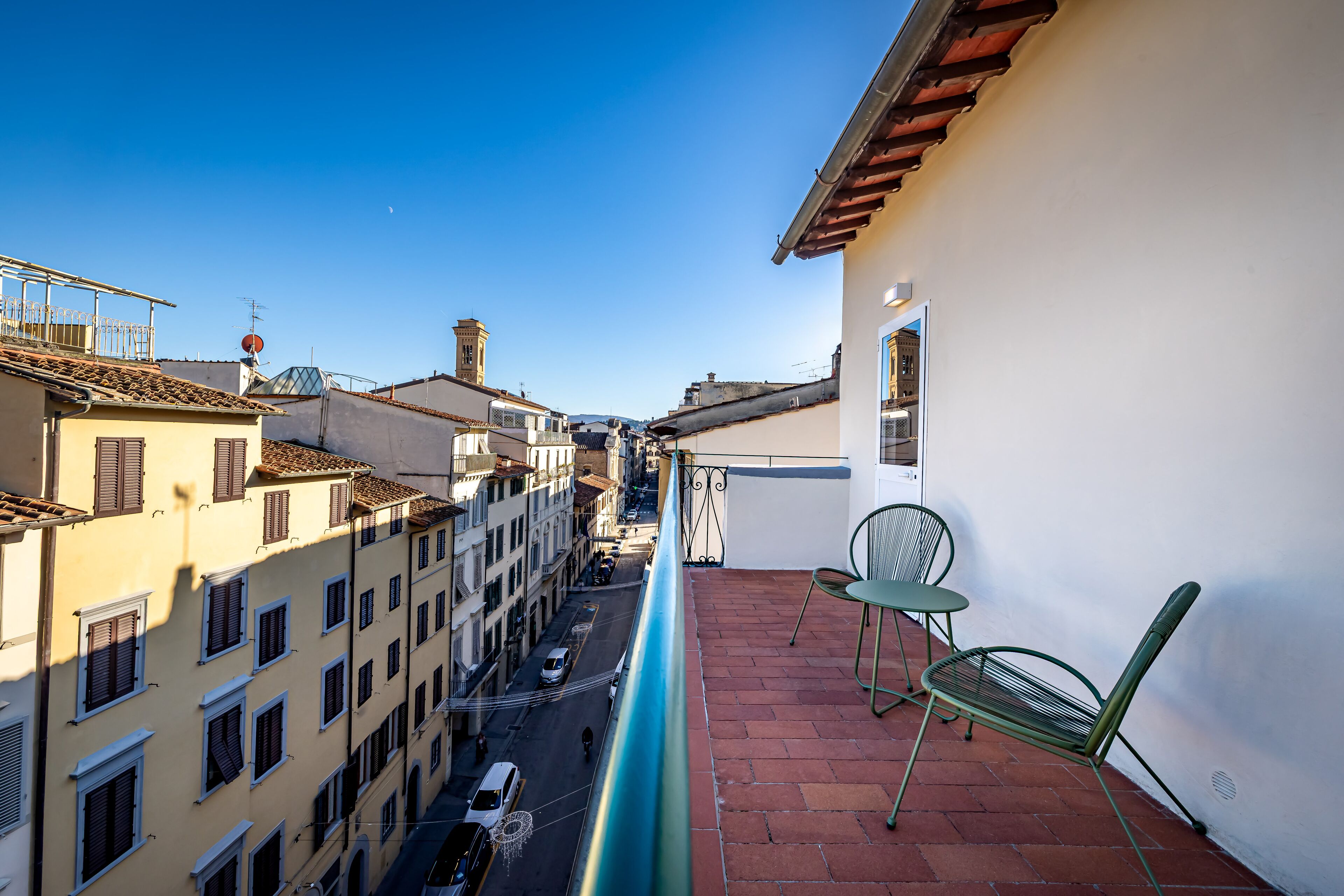 A narrow street with buildings on either side, a balcony with a chair, and a clear blue sky.