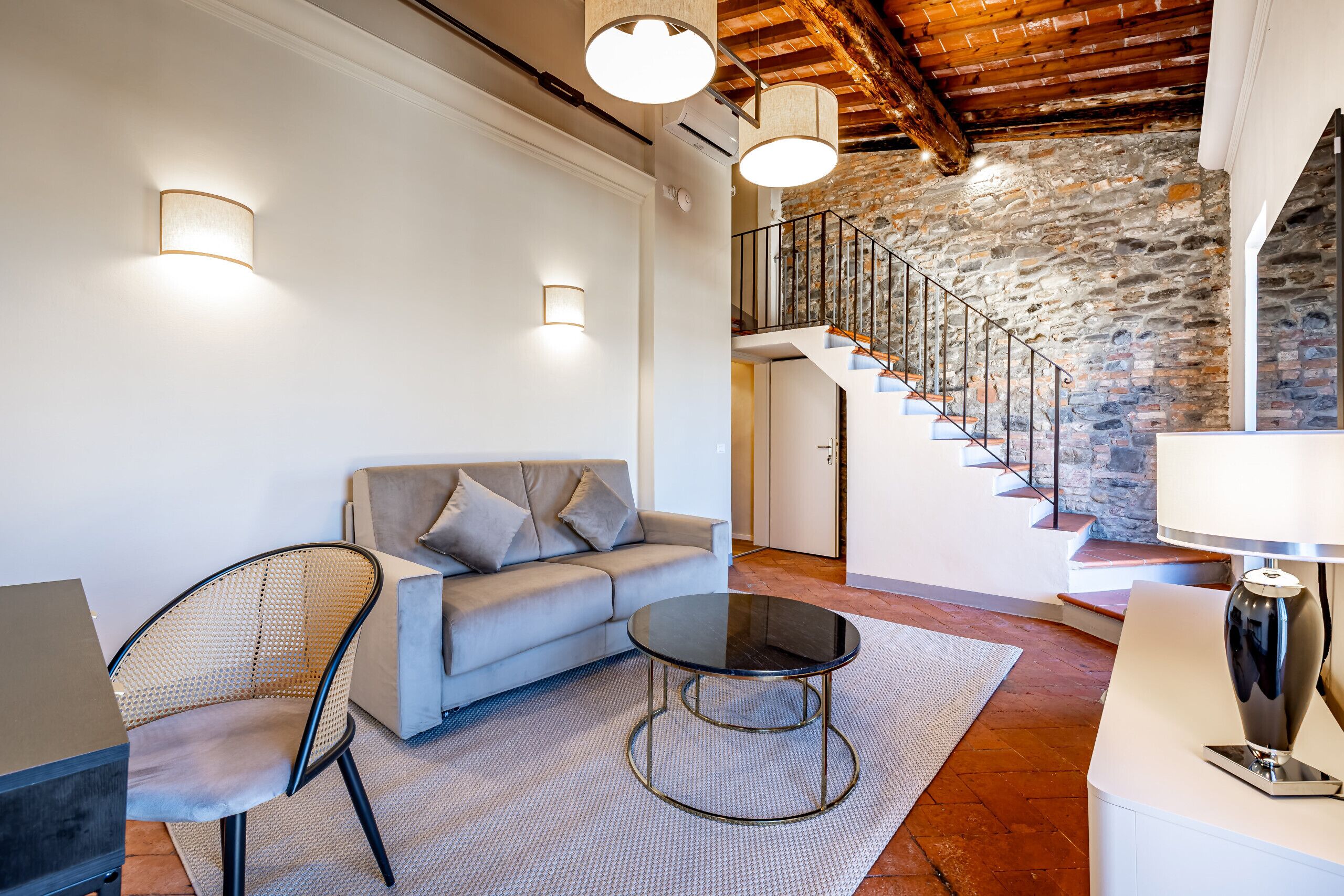 A modern living room with a sofa, a round table, a chair, a lamp, and a staircase with stone walls.