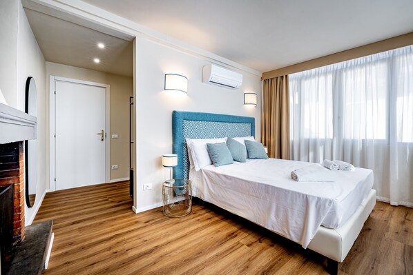 A modern hotel room with a wooden floor, a bed with white bedding and blue headboard, a small side table, and a window with sheer curtains. - YiD Four suites Ognissanti (Florence)