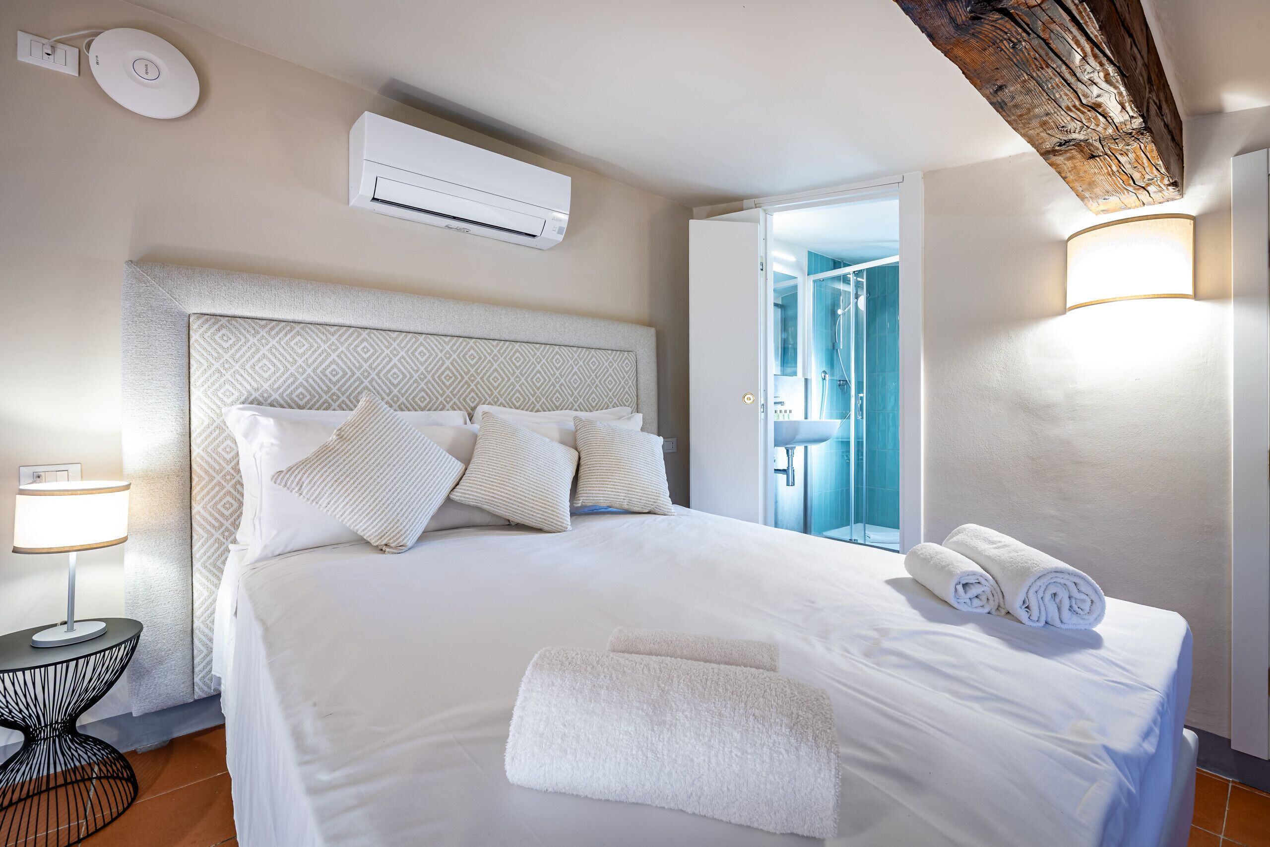A hotel room with a large bed, a bedside table with a lamp, an air conditioning unit, and a view of a bathroom.