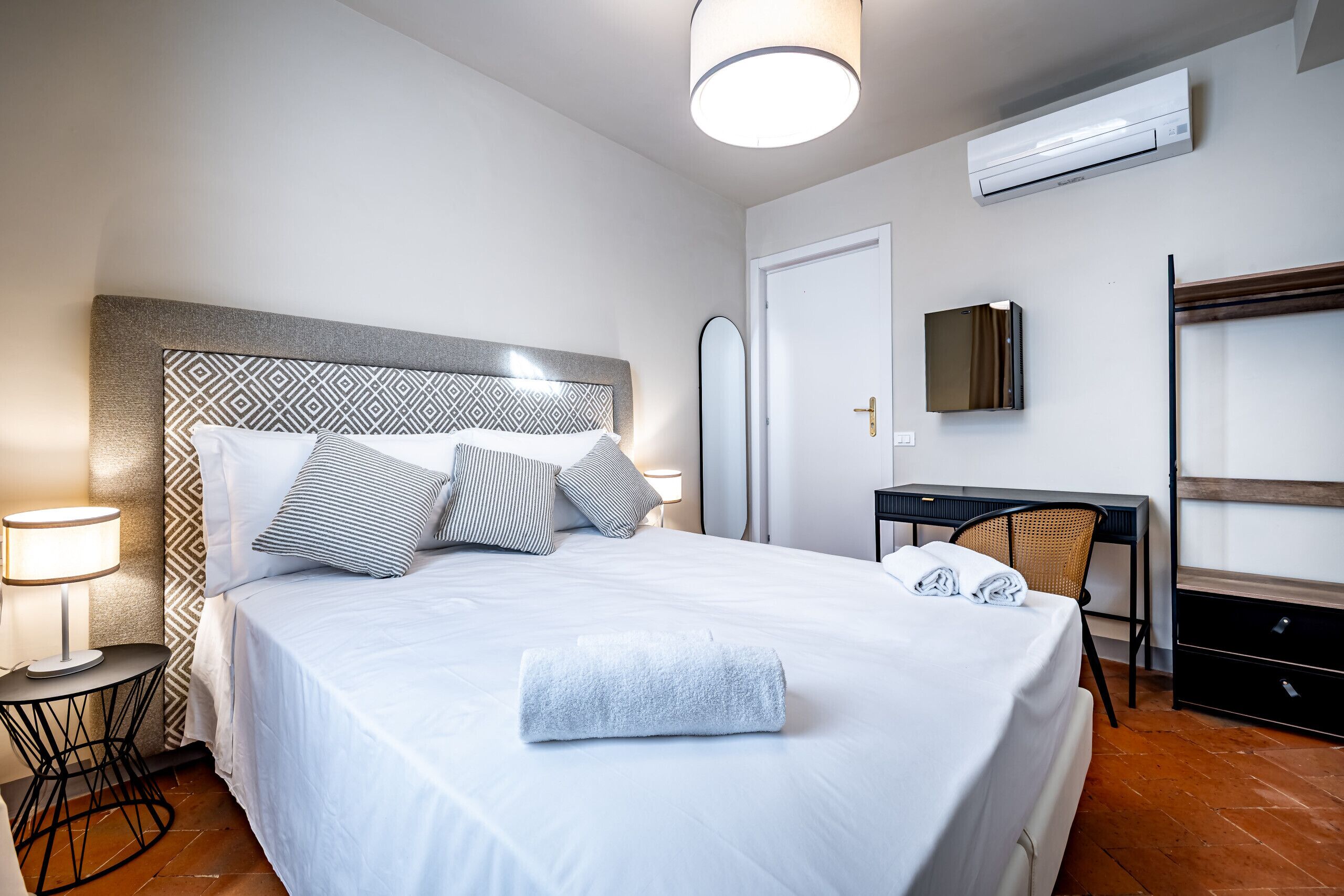 A neatly made bed with a white comforter and pillows, a bedside table with a lamp, a desk with a chair, and an air conditioning unit on the wall.