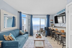 A living room with a sofa, coffee table, and bar stools. A large window offers a view of the ocean. - Oceanside Escape Condo w/ Ocean Views + Pools (Myrtle Beach)