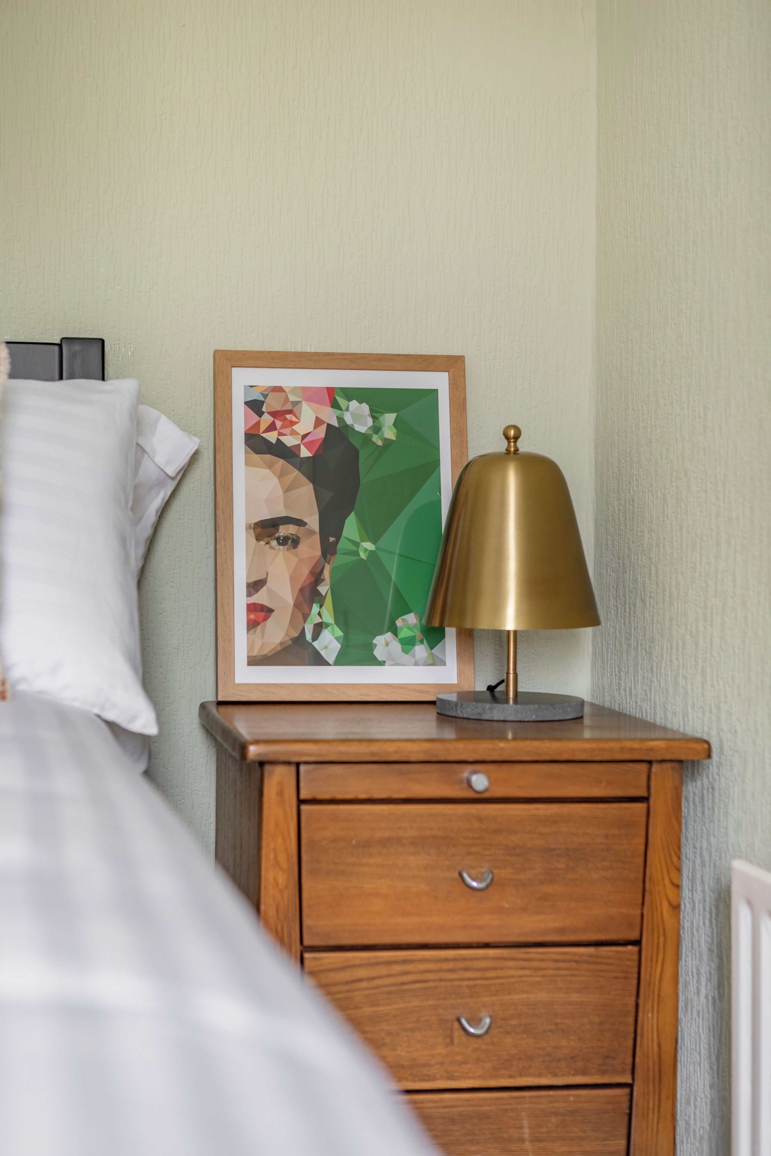 A neatly made bed with white linens, a wooden nightstand with a brass lamp, and a framed artwork on the wall.