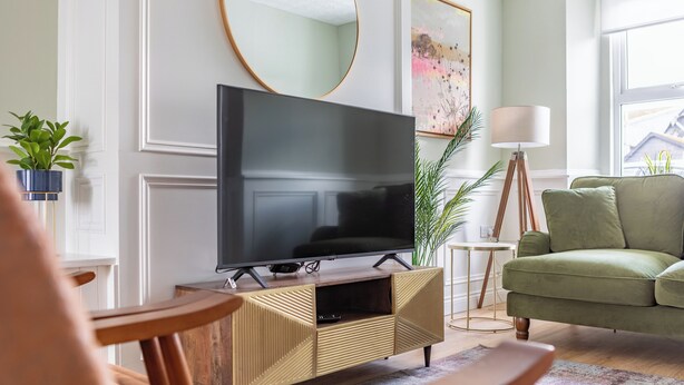 Ein Wohnzimmer mit Flachbildfernseher auf Holzfuß, einem grünen Sofa, einem kleinen runden Tisch und einer Stehlampe.