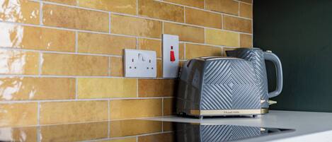 A kitchen with a tiled backsplash, a kettle, and a power outlet.
