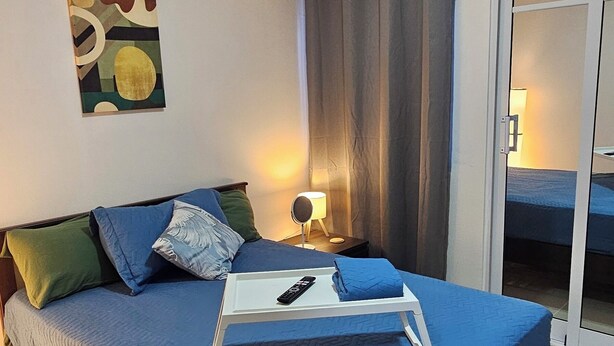 Una cama bien hecha con una colcha azul, un control remoto y una toalla doblada sobre una mesita de noche blanca.