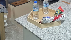 Una sala de estar moderna con un sofá gris, una mesa de centro con una bandeja que sostiene una botella de agua y aperitivos, y una zona de cocina con encimeras de granito.