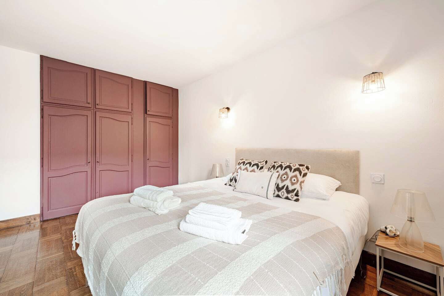 A neatly made bed with a striped bedspread, two pillows, and folded towels. There is a wooden nightstand with a lamp and a phone. The room has a large wardrobe with a pink door and a wall-mounted light fixture.