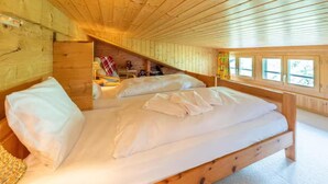 A wooden cabin bedroom with two beds, a small table, and a window with curtains. - Charming Swiss chalet with stunning mountain views. (Grächen)