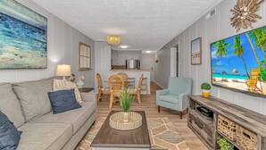 A modern living room with a sofa, coffee table, and TV. There is a dining area with chairs and a table. The room has a kitchen in the background. - I-2 2nd Floor Oceanfront with King Bed Suite (North Myrtle Beach)