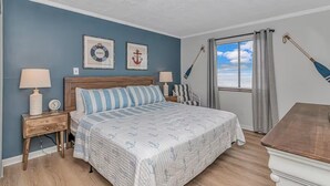 A bedroom with a bed, two nightstands, a wooden headboard, a window with curtains, and nautical-themed wall art. - I-2 2nd Floor Oceanfront with King Bed Suite (North Myrtle Beach)