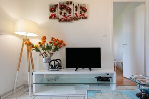 Una sala de estar con un televisor de pantalla plana, una lámpara, un jarrón con flores y una mesa de centro de vidrio.