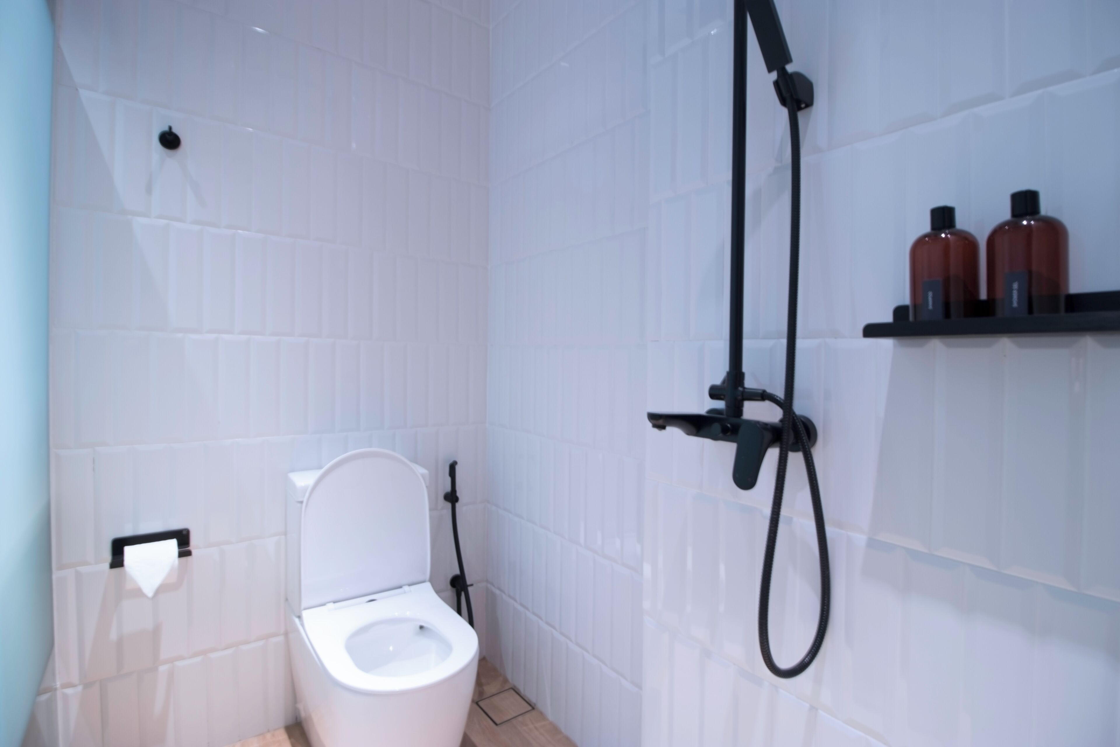 A bathroom with white tiled walls, a white toilet, a shower unit, and two shelves with bottles.