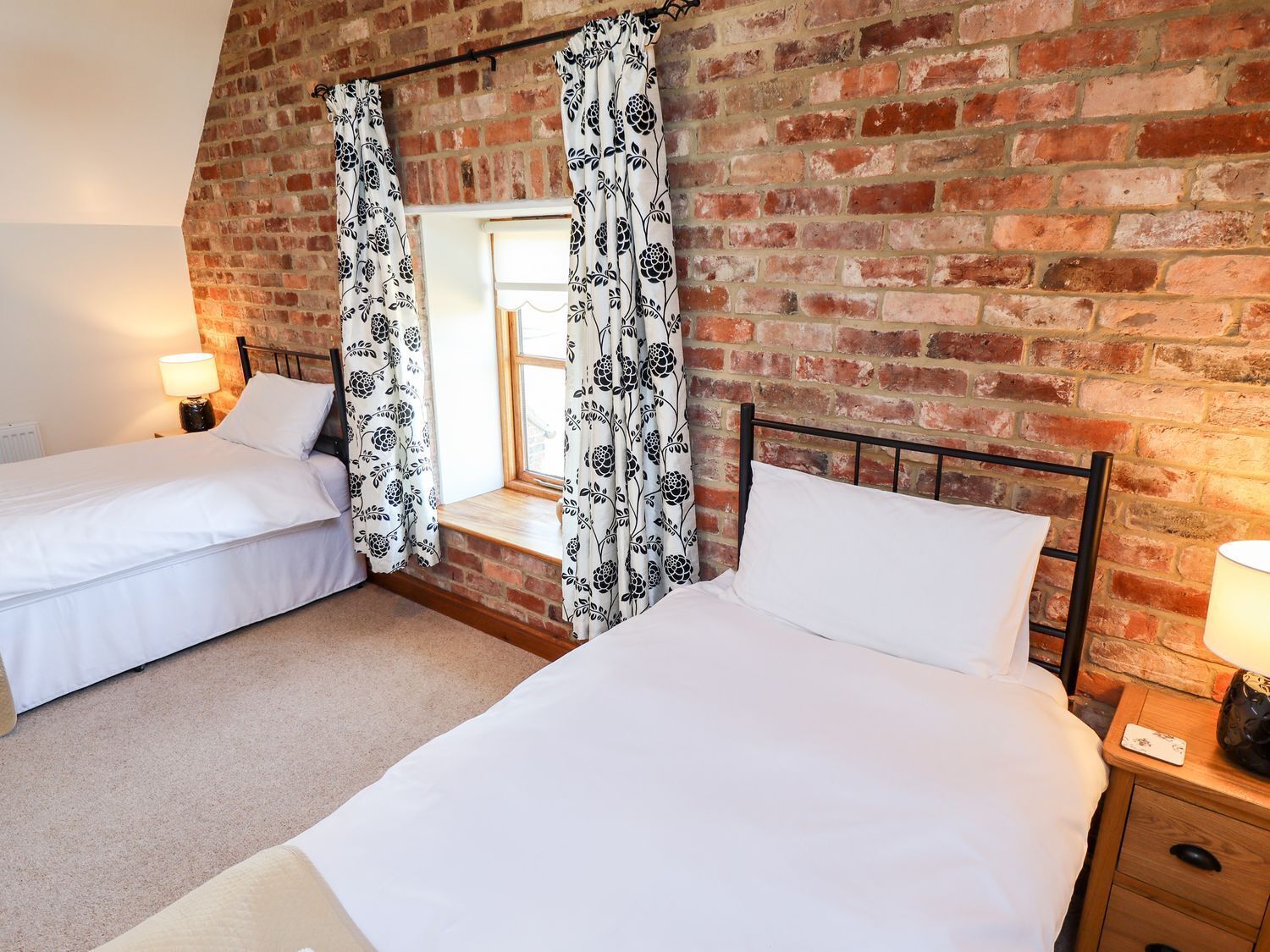 A bedroom with two beds, a brick wall, and a window with curtains.