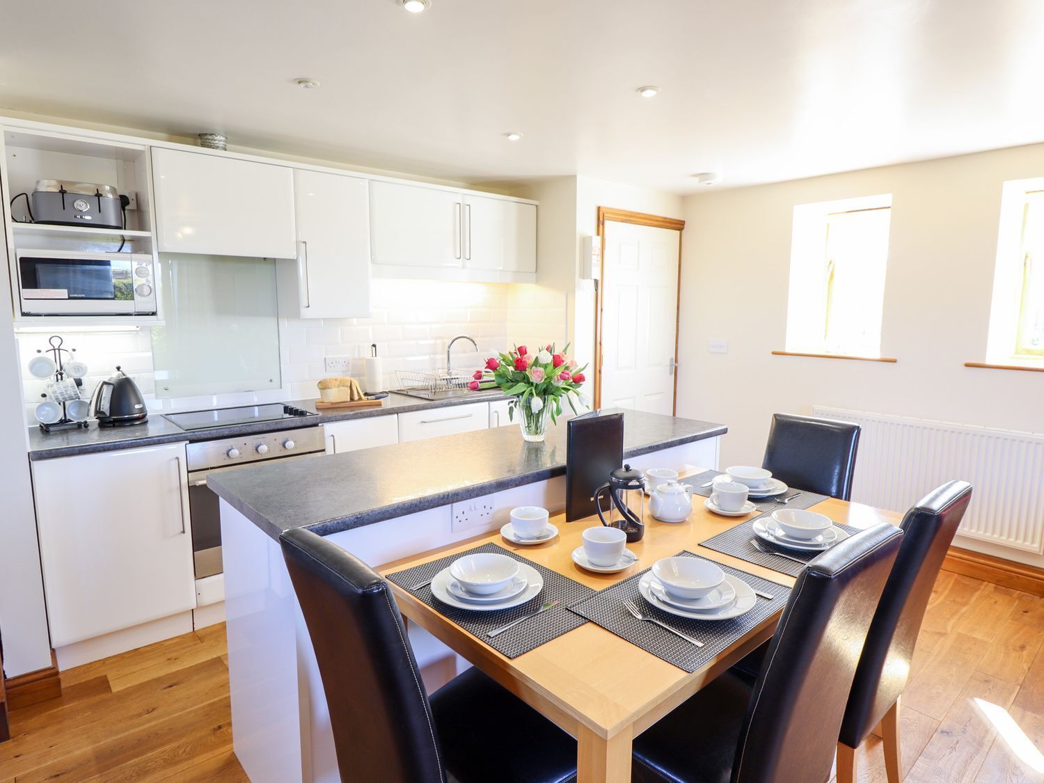 Una cocina moderna con armarios blancos, una zona de comedor con una mesa de madera dispuesta para cuatro comensales y un florero sobre la encimera.