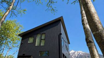 A modern two-story building with a dark exterior, surrounded by trees and a mountain backdrop.