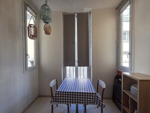 Un comedor con mantel a cuadros, dos sillas, un estante de madera con platos y una ventana con cortinas.