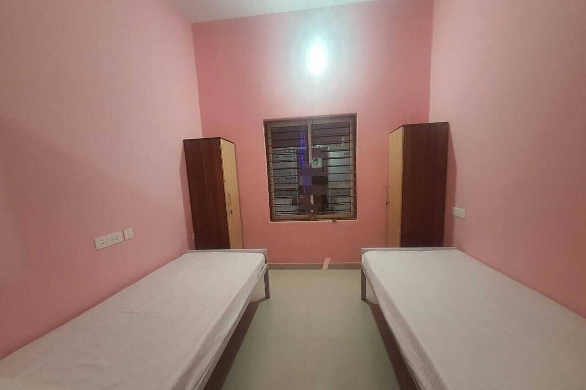 Une petite pièce aux murs roses, avec deux lits simples, une fenêtre grillagée et un luminaire au plafond.