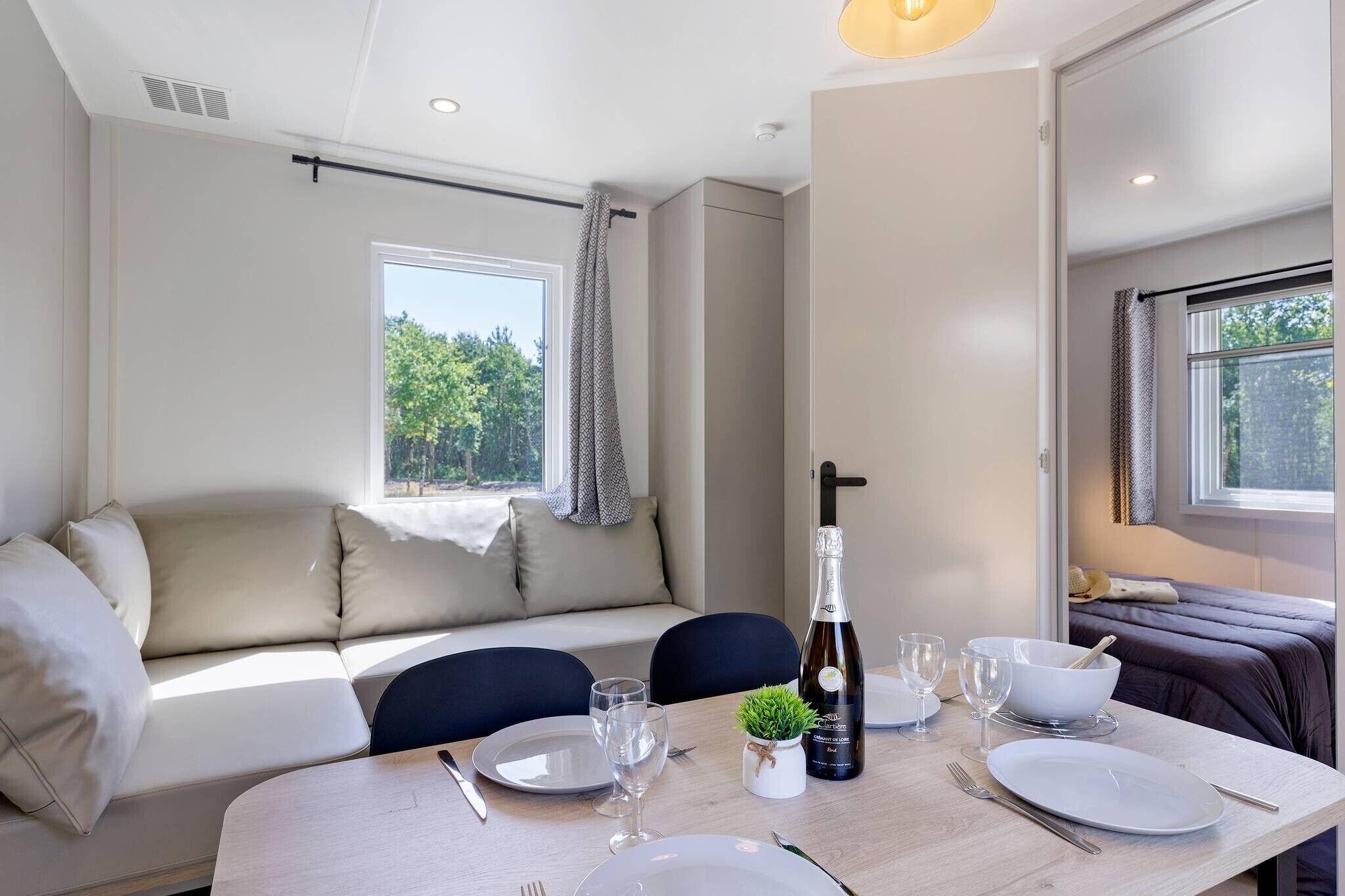 A room with a sofa, a dining table set with plates, glasses, and a bottle of champagne. There is a window with curtains and a view of greenery outside.