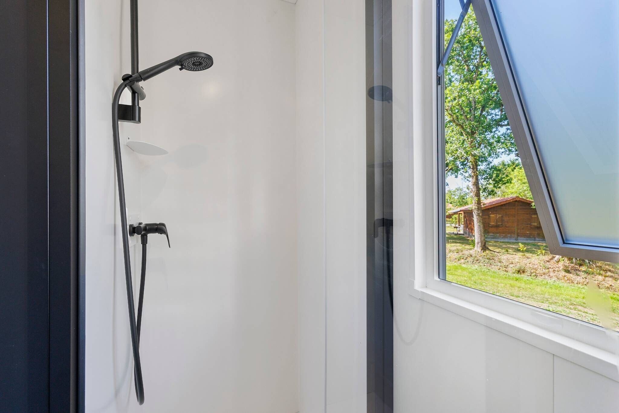 Een douche met een handdouche en een slang, een raam met uitzicht op een houten constructie en bomen, en een witte deur.
