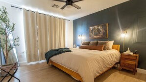 Ein Schlafzimmer mit einem Bett, Nachttischen, einem Deckenventilator, einem Fenster mit Vorhängen und einer Topfpflanze.