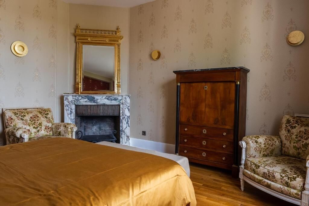 A bedroom with a large bed, a fireplace, a wooden wardrobe, and two upholstered chairs.