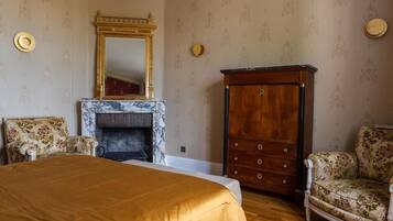 A bedroom with a large bed, a fireplace, a wooden wardrobe, and two upholstered chairs.