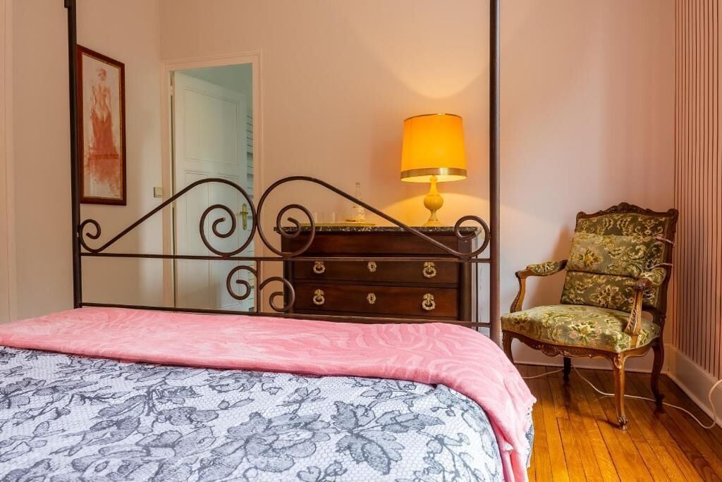 Een slaapkamer met een hemelbed, een dressoir met een lamp en een bloemen fauteuil.